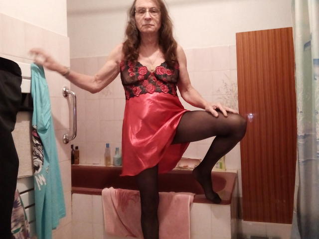 Hermeline, an older woman with long brown hair, wearing a red and black lace top with a red skirt, black stockings, and glasses, stands in a bathroom with one leg up.