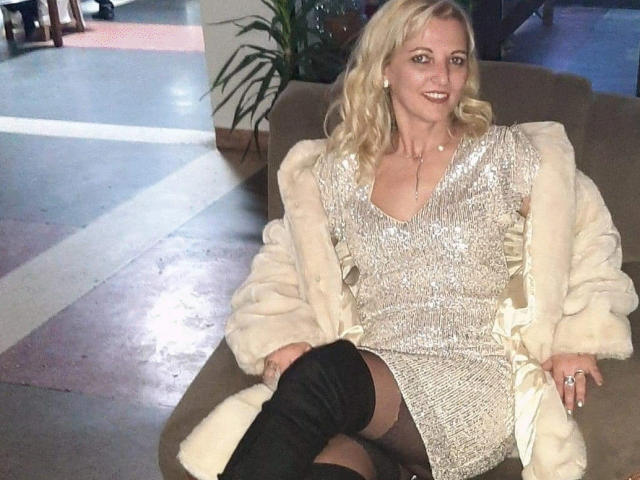 SexyCoco with blonde hair, wearing a sparkly gold dress, fur coat, and black thigh-high boots, sitting on a chair, smiling. Her tits are small, and she's sexy.