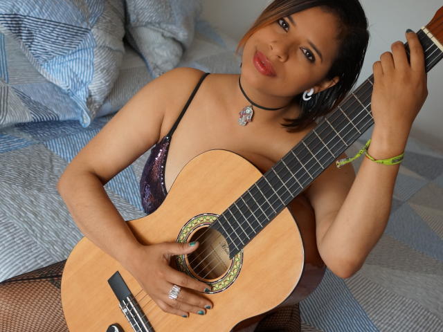 Karyn Black, con piel morena, cabello negro corto, lápiz labial rojo, vistiendo una blusa negra con lentejuelas, sosteniendo una guitarra acústica de madera, sentada en una cama con almohadas de estampado azul..
