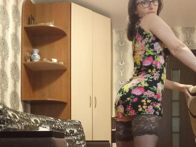 LaraxFish met een bril, een bloemenprint zwarte jurk en kanten hoge kniebedekkers, die haar achterwerk en zwarte kanten panty's laat zien, staand in een kamer met houten planken en witte kasten..