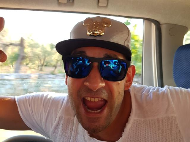 Un cammello con una camicia bianca, occhiali da sole blu riflettenti e un cappello bianco con un emblema dorato, con la bocca aperta in un'espressione sorpresa o eccitata, seduto in un'auto..