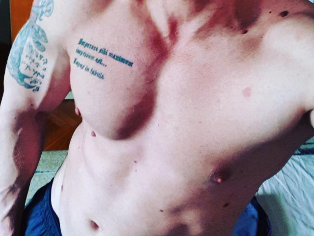 Het naakte, gespierde, bleke bovenlichaam van TonyLove69, met kleine tepels, tatoeages op de rechter schouder en borst, en een licht buikje, draagt donkere broek, in een goed verlichte kamer..