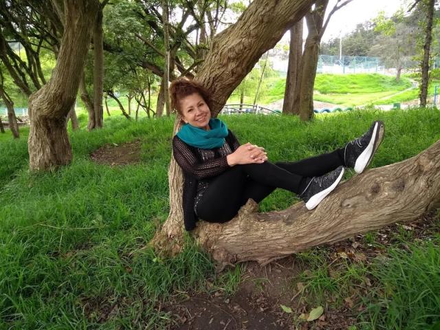 LadyLucky's ass is perched on a thick tree branch, wearing black pants and white sneakers, with a blue scarf, smiling, in a grassy park.