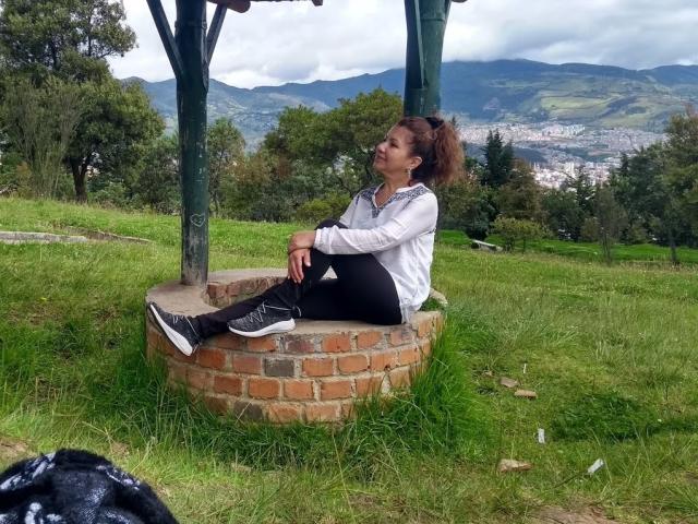 LadyLucky con una coda di cavallo marrone, una camicia bianca a maniche lunghe, pantaloni neri e scarpe da ginnastica nere, seduta su un cerchio di mattoni, in un'area erbosa, con pali di legno verdi, montagne e città sullo sfondo..