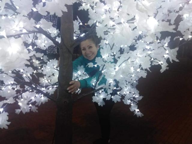 LadyLucky with short brown hair, smiling, wearing a teal jacket, hanging from a tree adorned with white paper flowers and glowing lights, standing on wooden floor.
