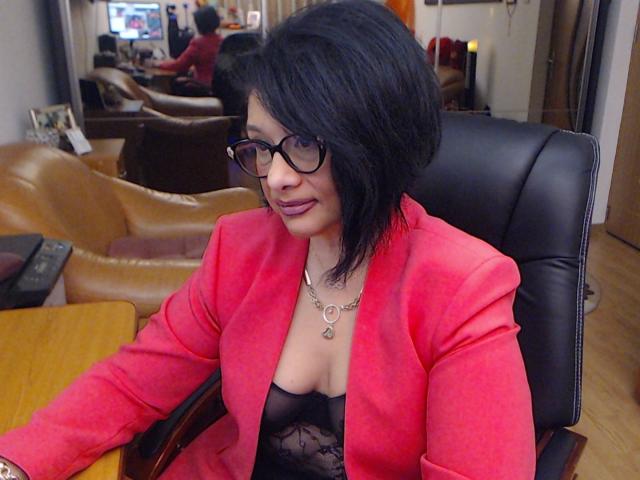 ClassybutNaughty with short black hair, glasses, and pink blazer, showing her black lace bra and small tits, sitting in a leather chair, smiling slightly.