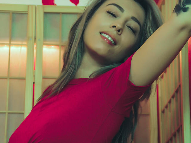 PoxyVibe with eyes closed, smiling, arms raised, wearing a red shirt, showing a tattoo on her right arm, blonde hair, red lipstick, and light skin.