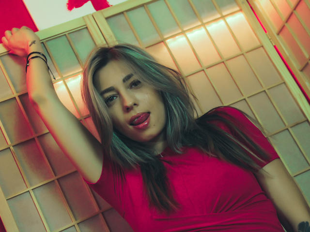PoxyVibe with blonde hair, red shirt, arm raised, tongue out, looking at camera. She's got a cute face, light skin, and a sexy vibe. Her tits are medium.