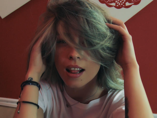 PoxyVibe with messy blonde hair, hands in hair, tongue slightly out, wearing white shirt, black bracelet on right wrist, looking slightly confused or annoyed.