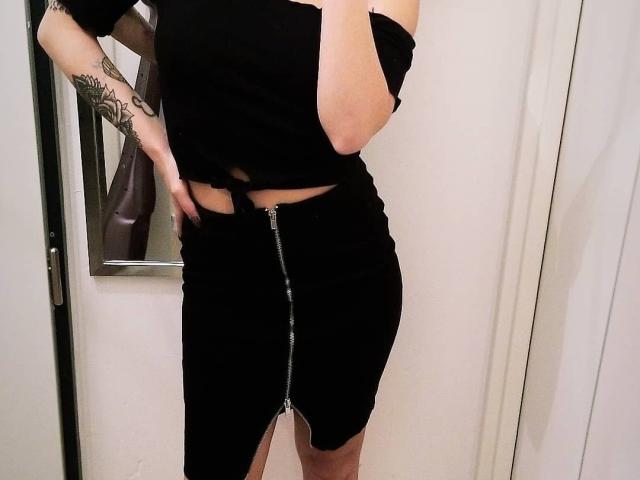 KassiKassou's black off-shoulder top, black skirt with silver zipper, and tattooed arm, showing a small cutout just above the skirt's zipper, in a mirror selfie.