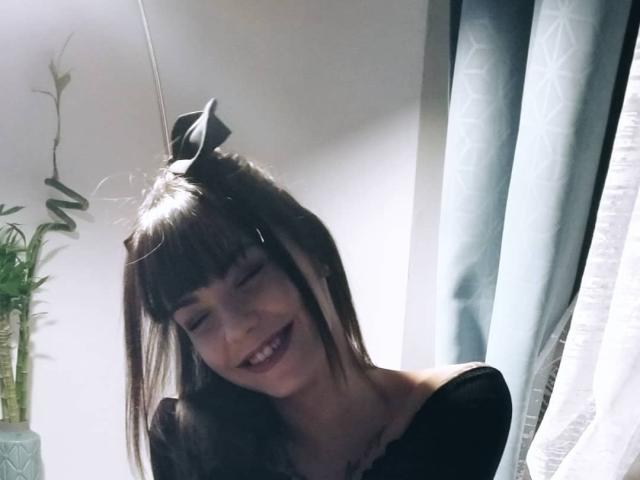 KassiKassou, con cabello negro, reflejos blancos y un lazo negro en la parte superior, sonriendo con los ojos cerrados, vestida con una blusa negra con un hombro descubierto, sosteniendo una prenda negra..