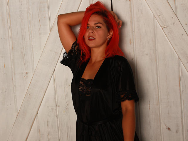 MelanyTessk with red hair, black lace dress, and black velvet robe, leaning against white wooden door, arm above head, looking seductive with a slight smile, full ass visible.