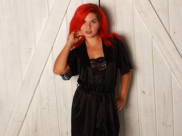 MelanyTessk with bright red hair, wearing a black lace top and satin robe, standing against white wooden doors, hand touching her hair, looking slightly to the side.
