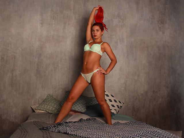 MelanyTessk with red hair, green lingerie, holding a red top, standing on a bed, hand on hip, arm raised, showing her tits and ass, in a studio.