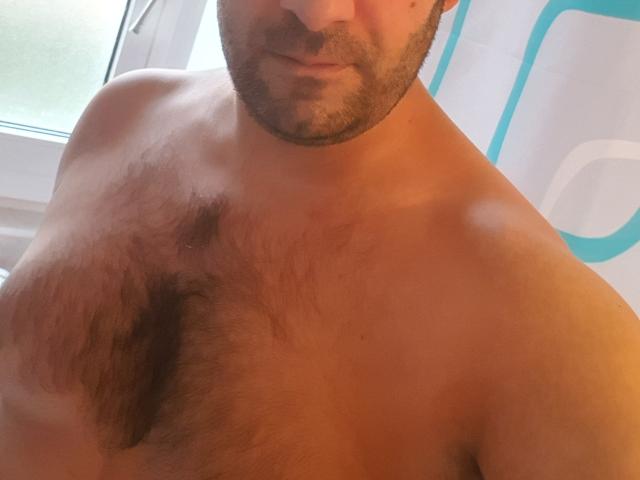 Camelhard zonder shirt, met een harige borst en baard, met een lichte glimlach, bovenlichaam zichtbaar, lichtbruine huid, raam op de achtergrond..