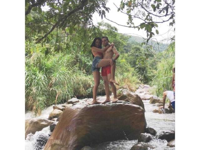 Stefany et Juan, debout sur un rocher dans un ruisseau, Stefany a les bras autour de la taille de Juan, tous les deux sans chemise, les seins de Stefany sont visibles, Juan porte un short rouge, et il y a une végétation verte autour..
