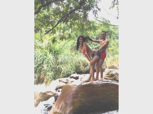 StefanyAndJuan stand on a rock, Stefany in a red bikini, Juan in red shorts, his hand on her ass, green foliage above, water flowing around their feet.