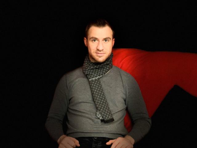 Un hombre atractivo con cabello castaño corto, vistiendo una camisa gris y una bufanda estampada, sentado frente a un sofá rojo, fondo negro, con las manos en el regazo, sonriendo ligeramente..
