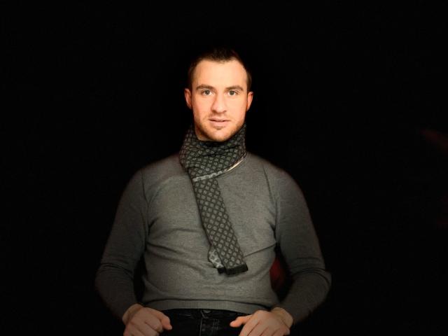 OneSexyGuy with a gray turtleneck, patterned scarf, and short brown hair, sitting against a dark background, hands on his lap, looking slightly to the side with a subtle smile.