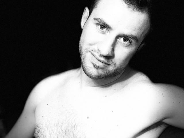 Zwart-wit foto van OneSexyGuy met een ontbloot bovenlijf, die de camera aankijkt met een lichte glimlach, donker haar, baard en een paar kleine moedervlekken op zijn borst..