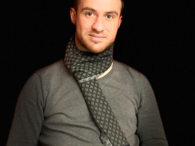 Un uomo attraente con una sciarpa a fantasia, che indossa una camicia grigia, ha un leggero sorriso, capelli castani corti e una leggera barba, su uno sfondo nero..