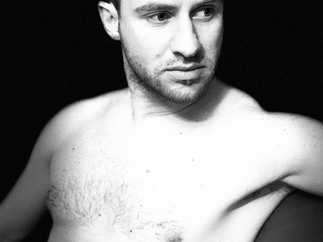 Shirtless OneSexyGuy with a serious face, short dark hair, light skin, slight beard, and chest hair, looking slightly to the right in a black-and-white photo.