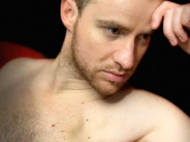 OneSexyGuy hat einen ernsten Gesichtsausdruck, helle blaue Augen und einen kurzen Bart. Er ist ohne Hemd, mit einer behaarten Brust, und legt sein Kinn in seine Hand..