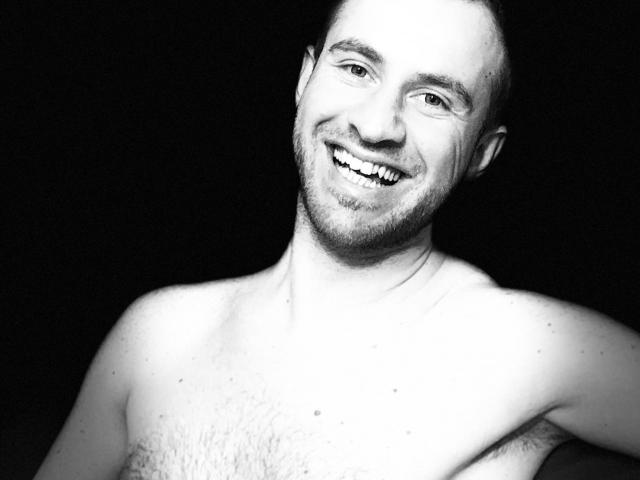 Zwart-wit foto van OneSexyGuy, zonder shirt, met een vrolijke glimlach, een korte baard en enigszins rommelig haar, waarbij een behaarde borst te zien is tegen een donkere achtergrond..