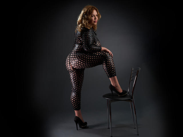 HornyyDiva con cabello rubio rizado, vestida con un traje de mallas negro transparente, tacones altos negros, y de pie con una pierna sobre una silla, mostrando su trasero..