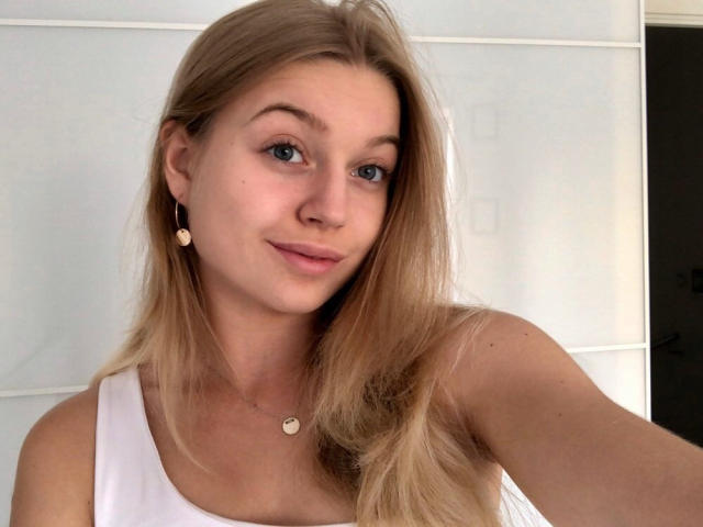 EricaLuck has blonde hair, blue eyes, and a slight smile. She's wearing a white tank top and small heart earrings. Her tits are visible but not the focus.
