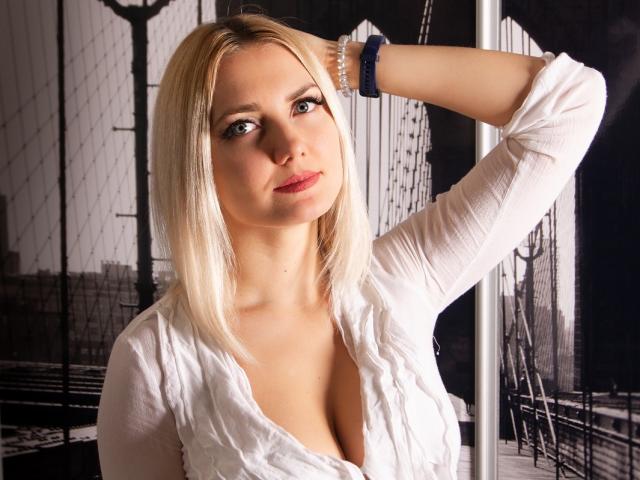 EliseONE has blonde hair, blue eyes, and a slight smile. She's wearing a white shirt, exposing her tits, with a blue bracelet on her right arm.