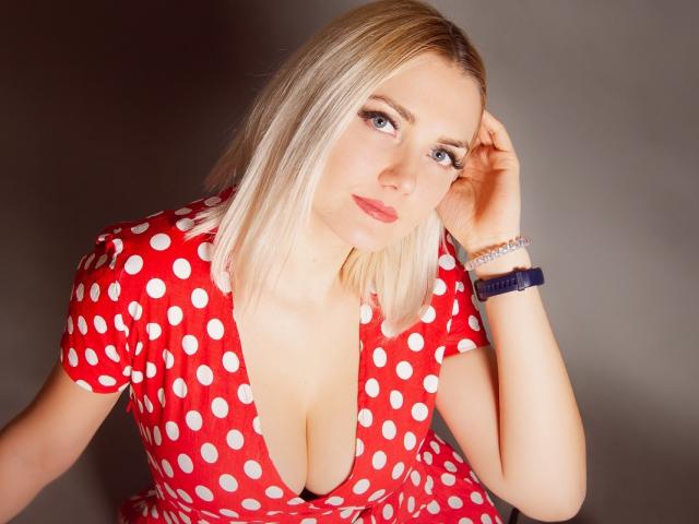 EliseONE with blonde hair, blue eyes, red polka dot dress, deep cleavage, pink lipstick, right hand touching head, blue bracelet, looking at camera, gray background.