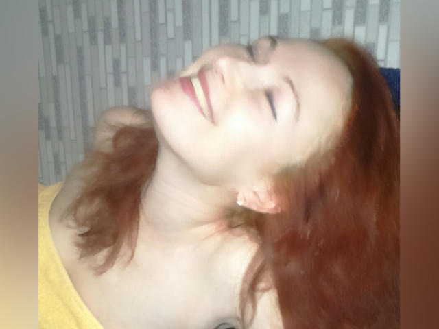MarleneDavies with red hair, eyes closed, smiling, head tilted back, wearing a yellow top, showing her neck and earrings, soft light, slightly blurry, looking happy and relaxed.