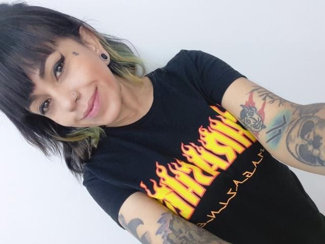 Pamela: Zwart haar met groene punten, neusring, tatoeages op de armen, zwart "Firewind" T-shirt, oorbel piercings, lachend, gemiddelde borsten, lichte huid en groene ogen..