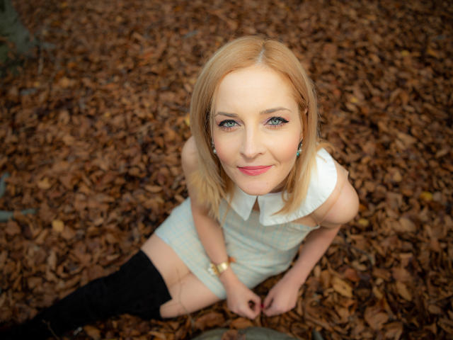 SoleildeNuit has blonde hair, blue eyes, and a cute smile, wearing a white dress and black boots, sitting on brown leaves, looking up with a slight blush.