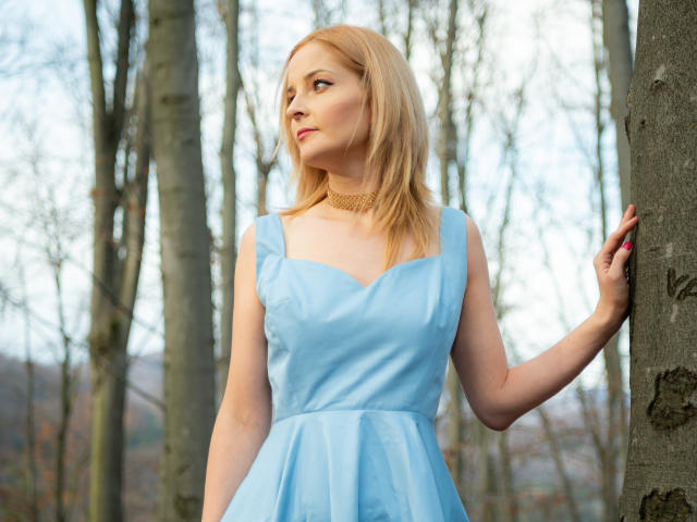 SoleildeNuit, con cabello rubio, un vestido azul claro y un collar, está de pie junto a un árbol, con una mano apoyada en el tronco, mirando hacia un lado. Se pueden ver los senos debajo del vestido, con un fondo de bosque..