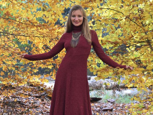 IampreciousLover, com cabelo loiro e um sorriso, está em um vestido vermelho, com os braços abertos, usando um colar grosso, cercada por folhas amarelas de outono. Seus seios são pequenos, ela é fofa..