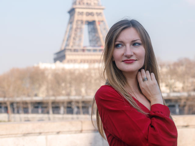 IampreciousLover com cabelos castanhos longos, vestido vermelho, sorriso discreto, olhos azuis, segurando a mão com um anel, Torre Eiffel desfocada ao fundo, luz suave, leve rubor nas bochechas..