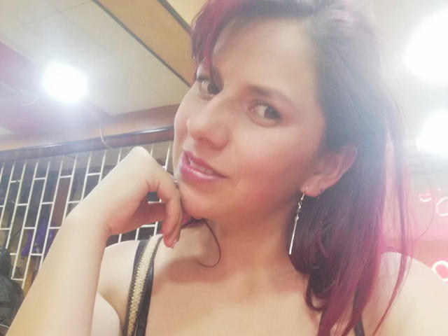 ScarletSexxy heeft rood geverfd haar, roze lippen en kijkt met een lichte glimlach naar de camera. Ze draagt een zwarte band en hangende oorbellen, en heeft haar kin rustend..