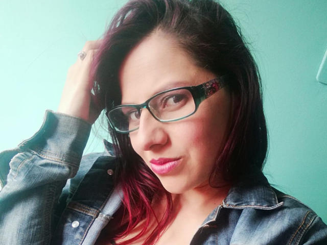 ScarletSexxy mit Brille, rosa Lippenstift und dunkelrotem Haar, trägt eine Jeansjacke, hat eine Hand im Haar und schaut in die Kamera, leicht lächelnd. Ihre Brüste sind nicht sichtbar..