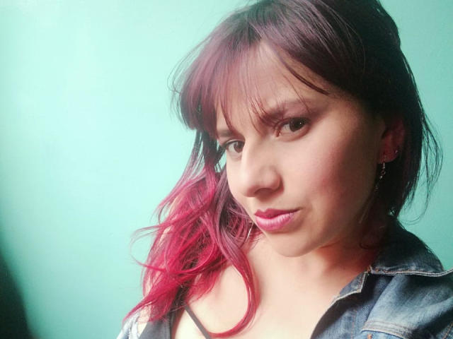 ScarletSexxy heeft roze haar, een pony en een denimjas, met roze lippenstift en een lichte frons, en kijkt met een schattige, sexy uitdrukking in de camera..