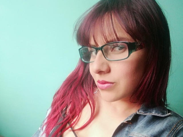 ScarletSexxy, con i capelli rosso-rosa, gli occhiali, la giacca di jeans, il rossetto rosa e un leggero sorriso, guarda di lato. Ha un aspetto carino, da intellettuale e sexy..