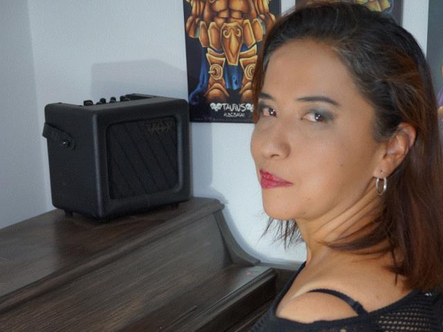 PaolaLady a la peau marron moyen, un haut en maille noire, du rouge à lèvres et des cheveux bruns. Elle se tient à côté d'une enceinte Rockbox noire sur une table en bois..