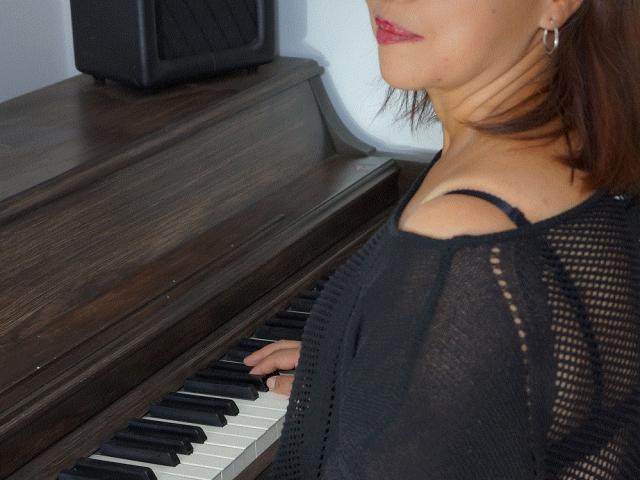 Paola, uma mulher de cabelos castanhos, batom vermelho e brincos de argola, tocando um piano de madeira escura, vestindo uma blusa de malha preta, mostrando seu ombro nu e seios pequenos..