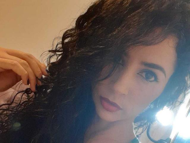 SexyKonnyX hat lockiges, schwarzes Haar, braune Augen und pinken Lippenstift. Sie berührt mit einer Hand ihr Haar, blickt in die Kamera und das Selfie ist gut beleuchtet, aber leicht unscharf..