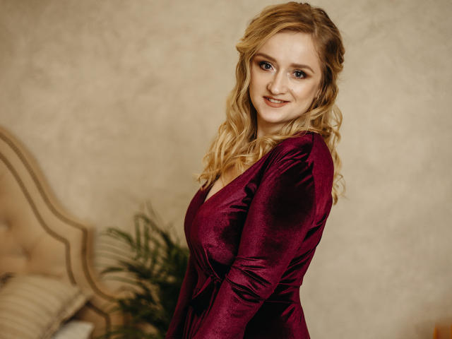 OhMollyHoney, blonde, wavy hair, red velvet dress, smiling, looking over shoulder, medium tits, soft lighting, bedroom, potted plant, tufted headboard, subtle makeup, cute, sexy.