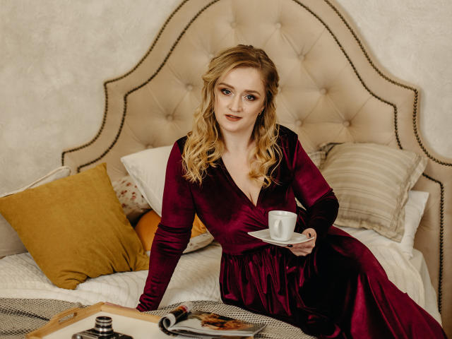 OhMollyHoney with blonde hair, wearing a deep red velvet dress, sitting on a bed holding a white cup, her tits slightly visible, surrounded by pillows and a tray.