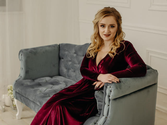 OhMollyHoney in a deep red velvet dress, sitting on a blue tufted sofa, with blonde wavy hair and a slight smile, hands resting on the sofa's arm.