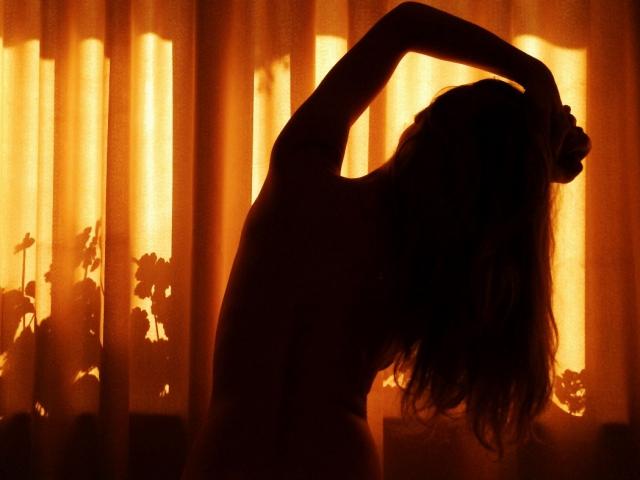 Silueta de iampreciousLover con los brazos levantados, de espaldas a la cámara, en una habitación oscura, con una luz naranja brillante que entra por la ventana, mostrando un cuerpo sexy, desnudo, curvilíneo, con glúteos prominentes y cabello largo..