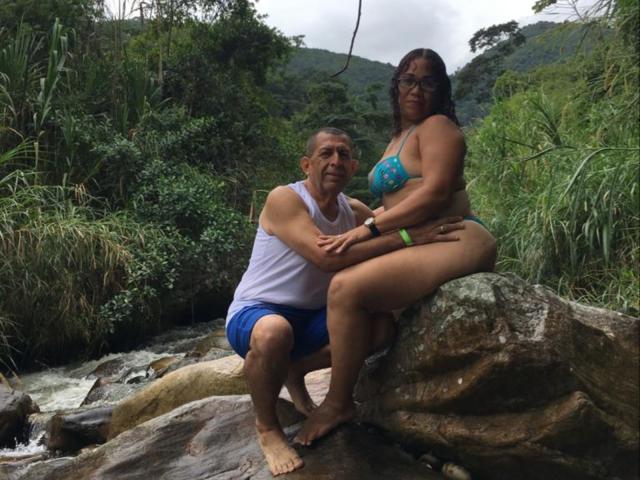 PamelaYDiego with big ass in blue bikini, sitting on rock, next to middle-aged guy in white tank top and blue shorts, by rocky stream in forest.